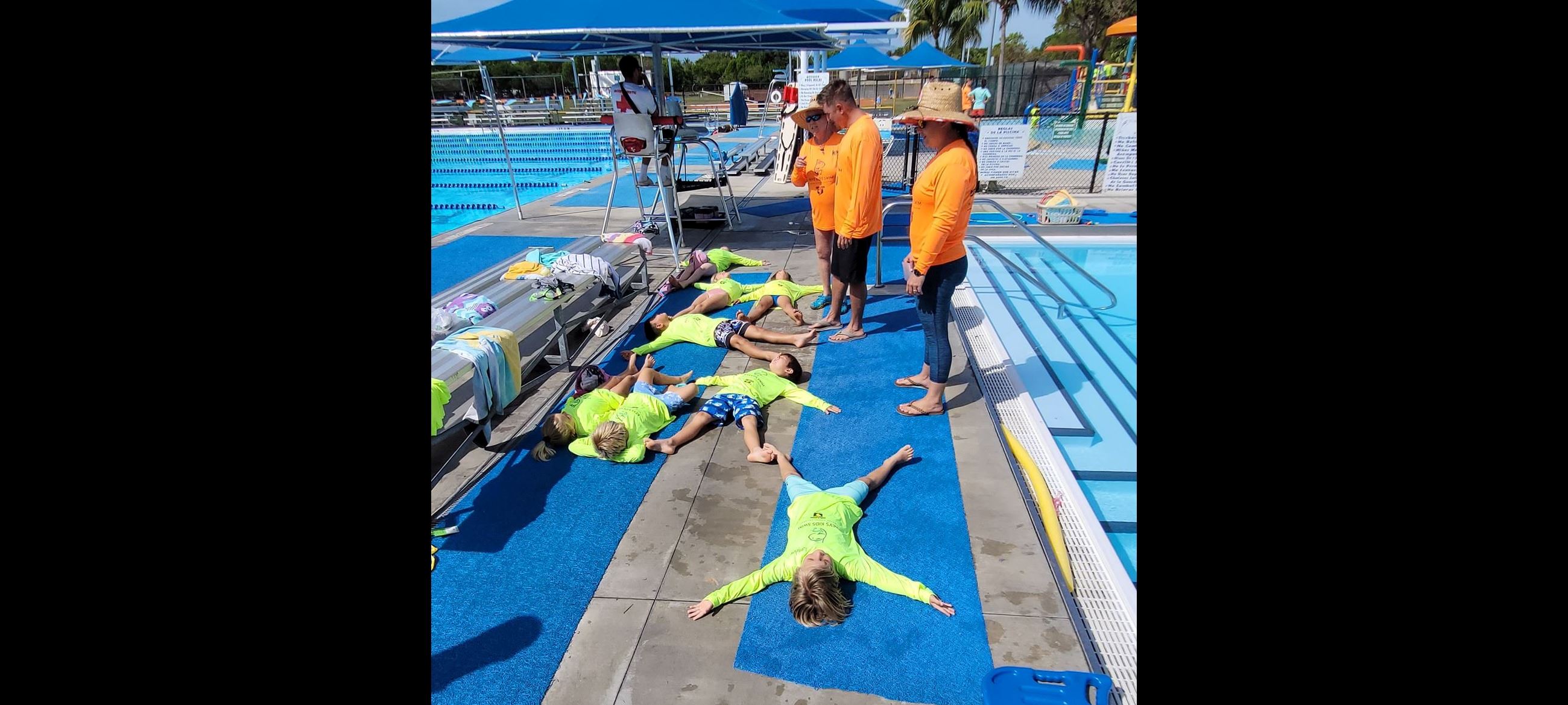 3 instructors in orange shirts stand over kids in yellow shirts lying on pool deck learning to float